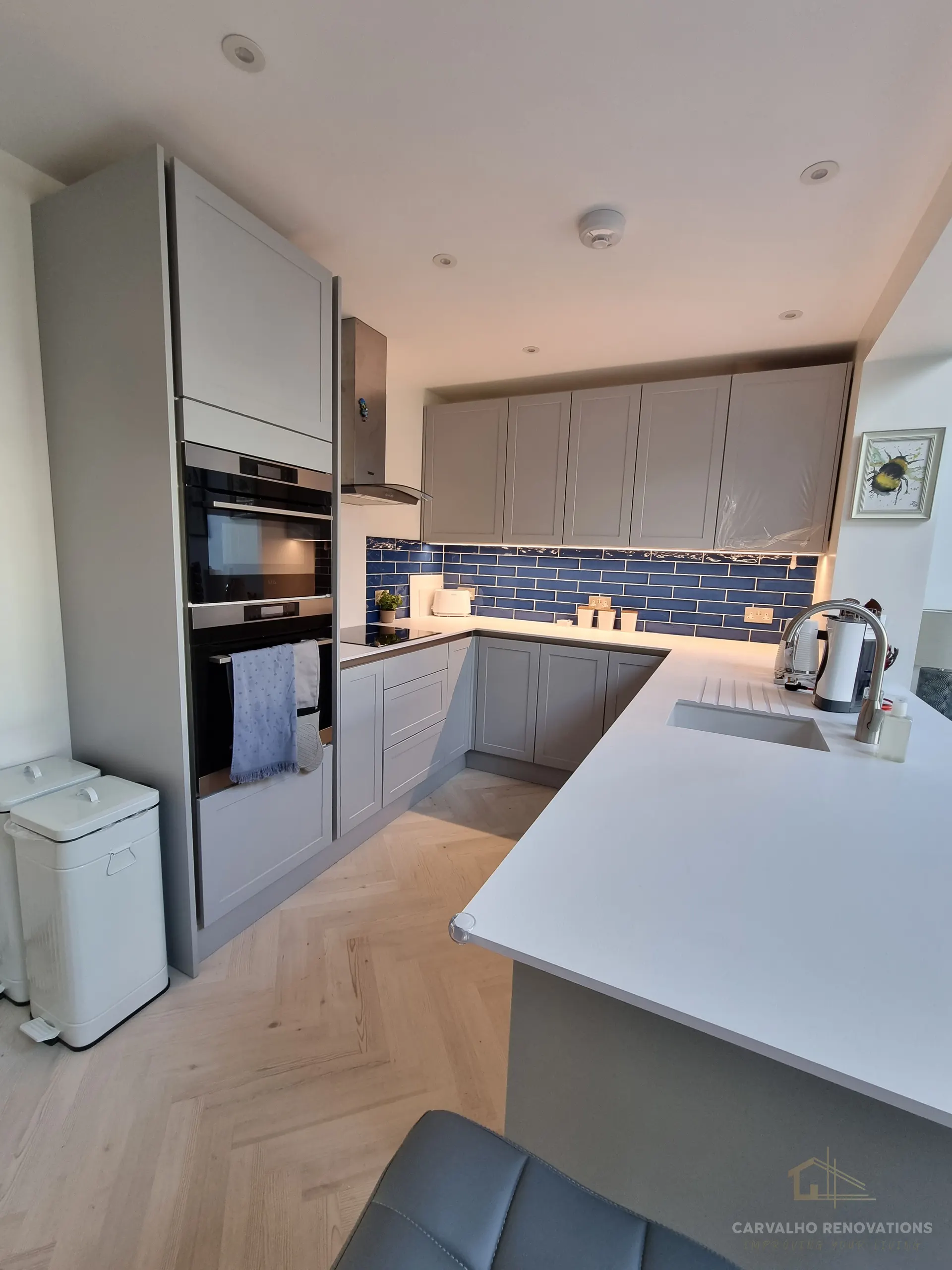 Grey Shaker kitchen in a U-shape with a blue metro-tile splashback, white worktops, built-in double oven and a peninsula with an inset sink.