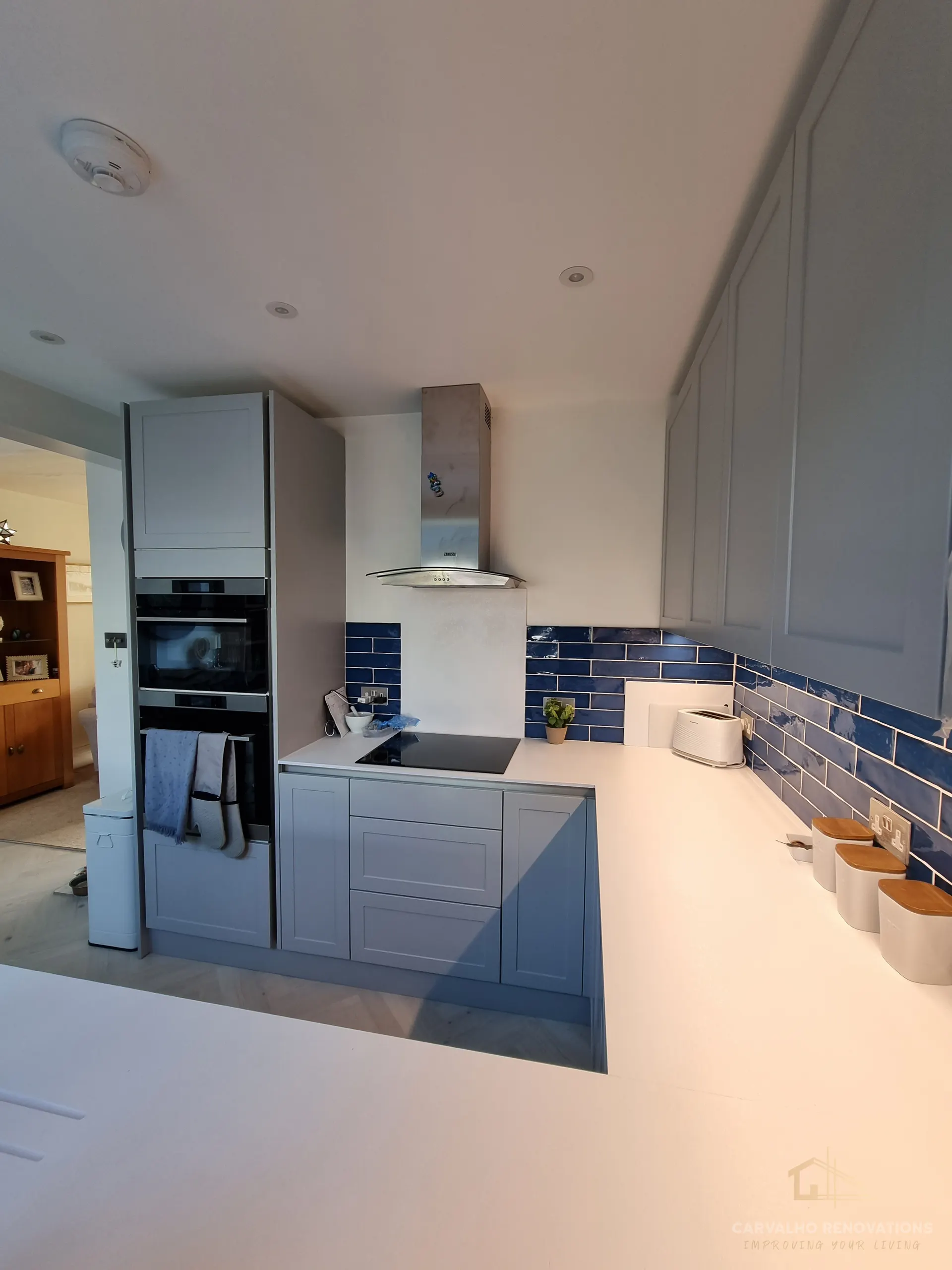 Induction hob beneath a stainless steel chimney extractor, with a blue metro-tile splashback and white worktop.