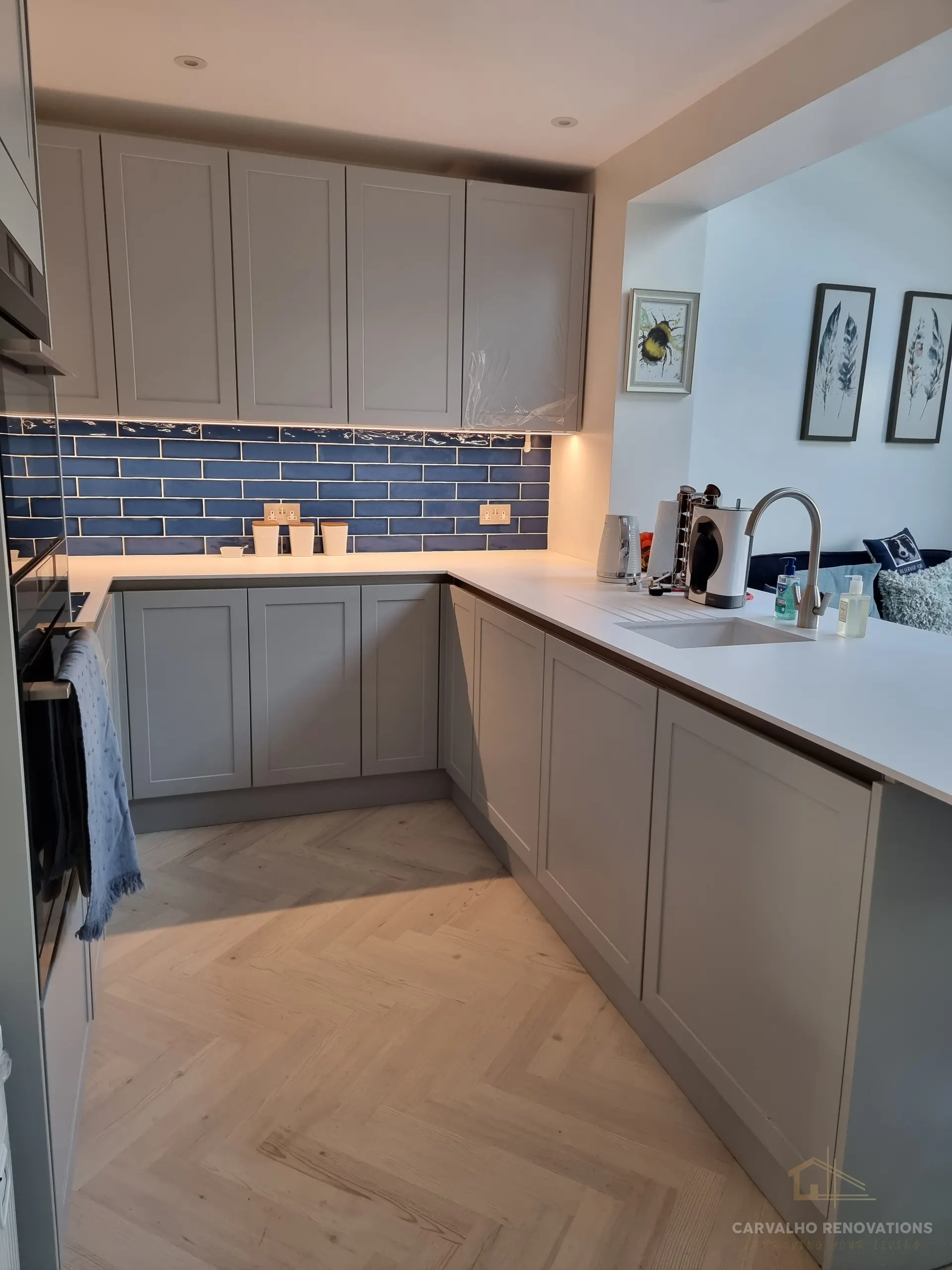 Corner of the grey kitchen showing the blue metro-tile splashback, under-cabinet lighting, white worktop and an undermount sink.