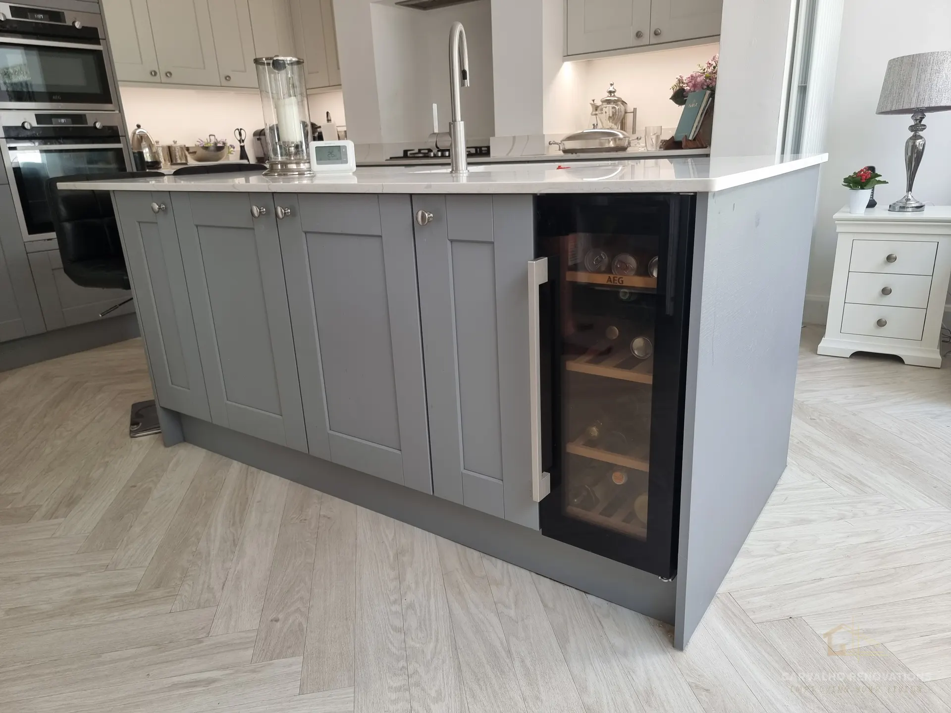 Grey Shaker kitchen island with a white quartz-effect worktop, brushed steel pull-out tap and a built-in wine cooler, on herringbone flooring.