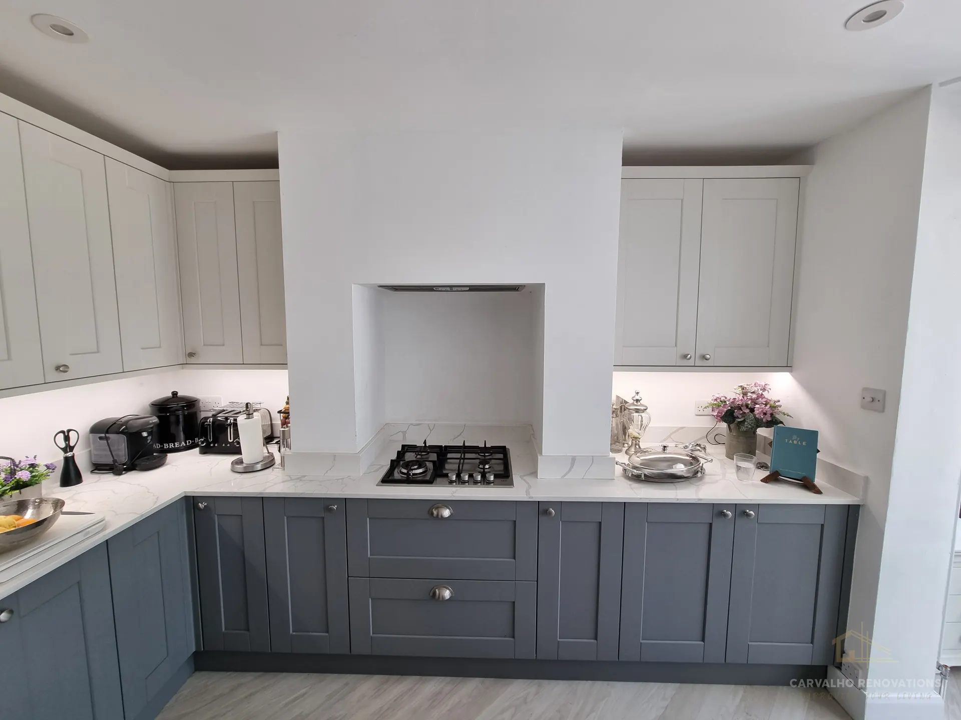 Wider view of the kitchen: light wall cabinets above dark grey base units, a gas hob beneath a recessed extractor, and under-cabinet lighting.